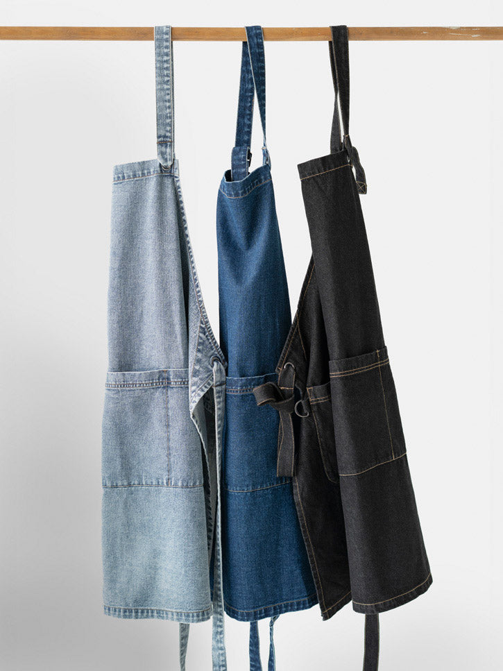 Biz Collection - Three denim aprons in light blue, dark blue, and black hang on a rack.