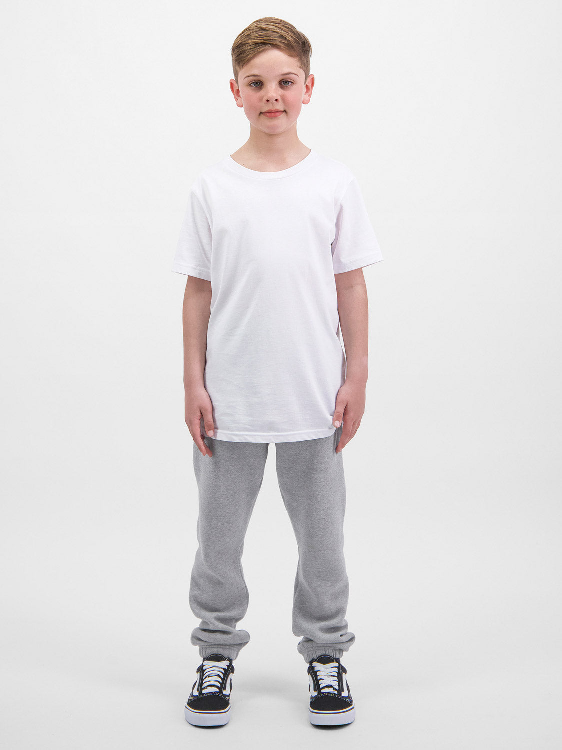 A young boy stands wearing grey Kids Basis Track Pants with a white t-shirt and black sneakers.