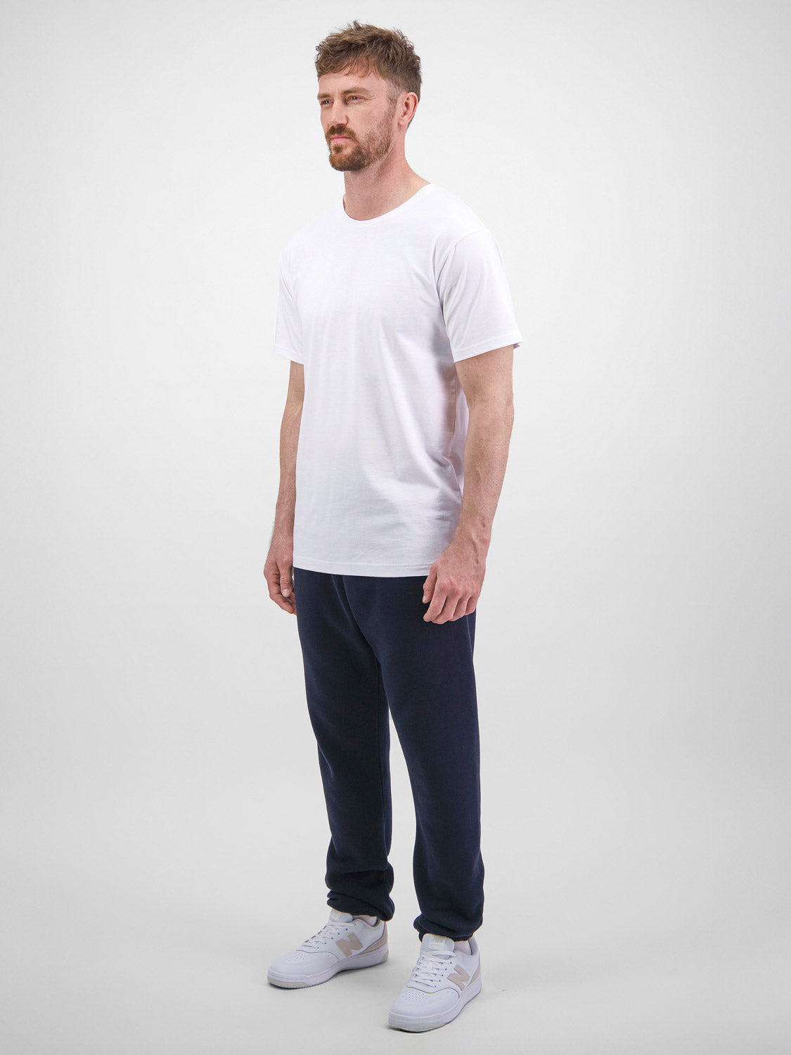 A man stands wearing navy blue Mens Basis Track Pants with a white t-shirt and sneakers.