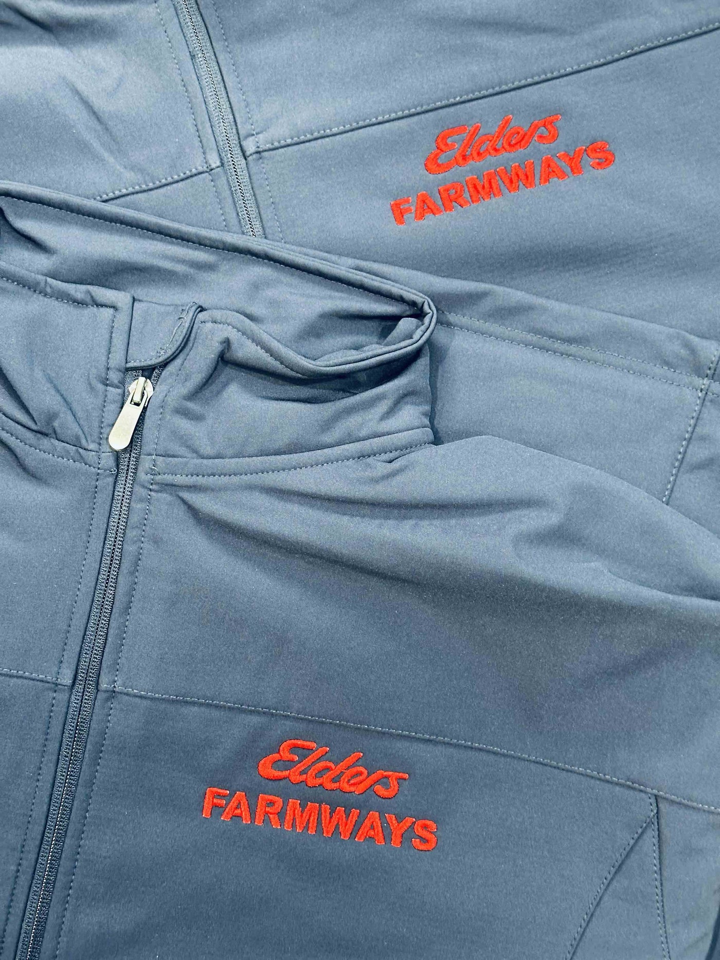 Close-up of a blue zippered jacket or sweatshirt with red embroidered 'Elders FARMWAYS' text on the chest and upper pocket area.