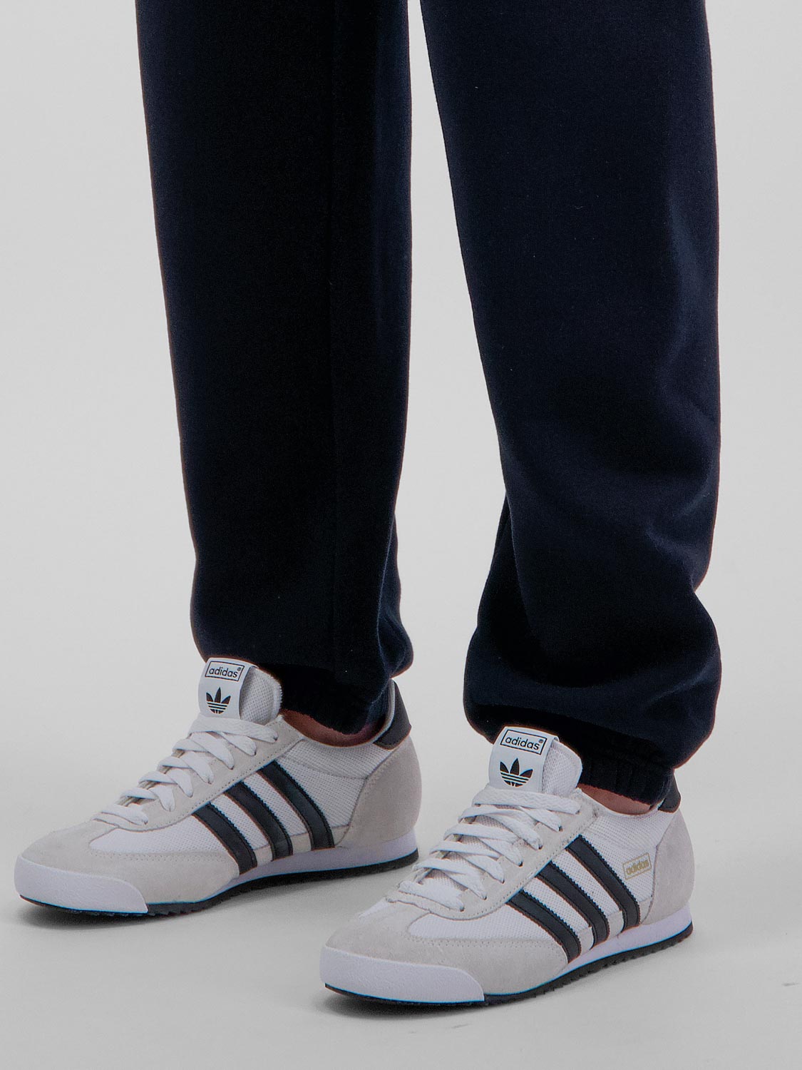 A person wearing navy blue Basis Track Pants with elastic cuffs and white Adidas sneakers.