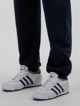 A person wearing navy blue Basis Track Pants with elastic cuffs and white Adidas sneakers.