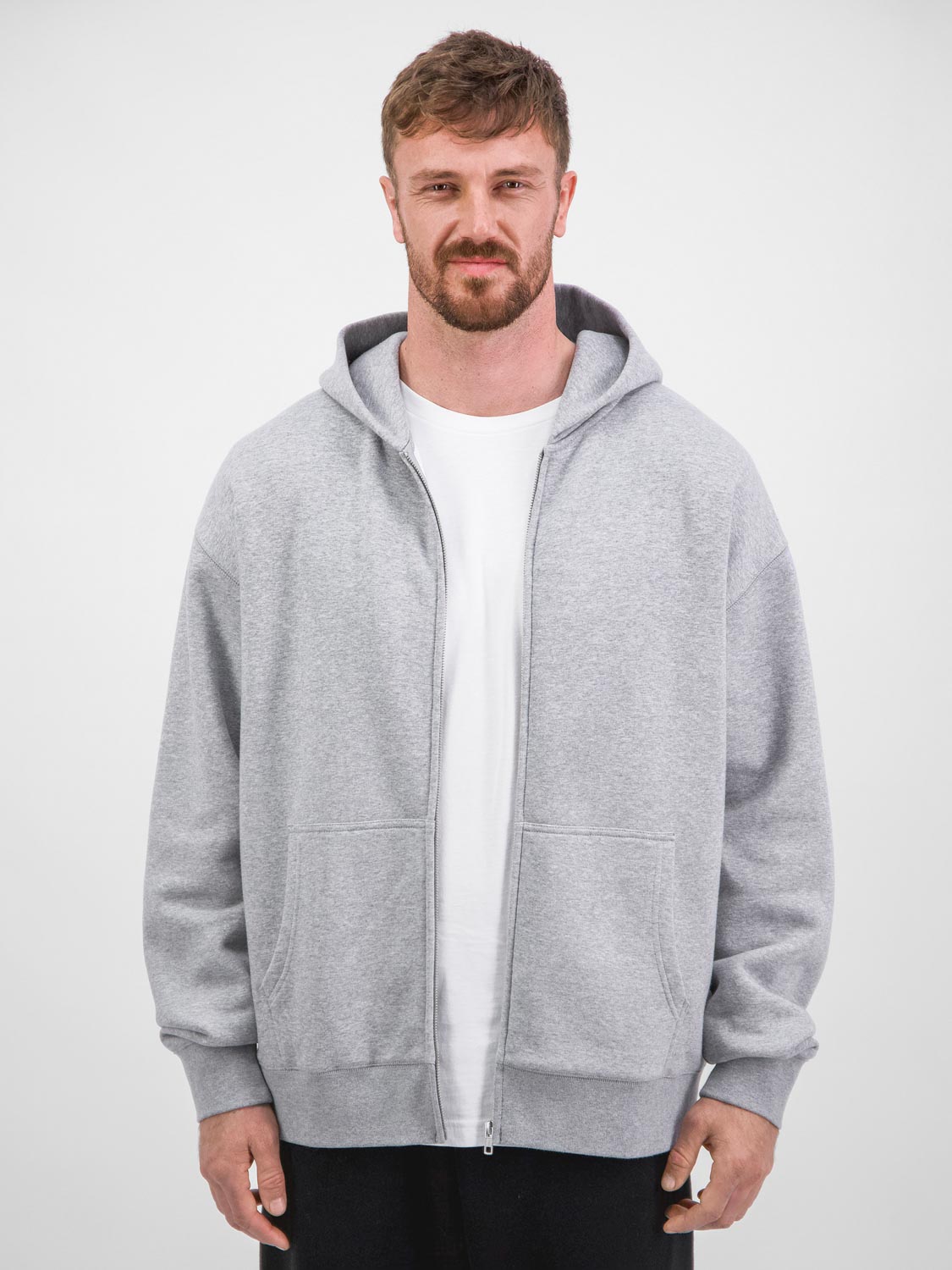 A man wears a grey zip-up hoodie with a white t-shirt and black pants against a white background.